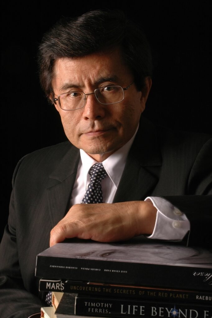homem de meia idade, vestido com terno preto, camisa branca e gravata escura, braço apoiado sobre livros empilhados
