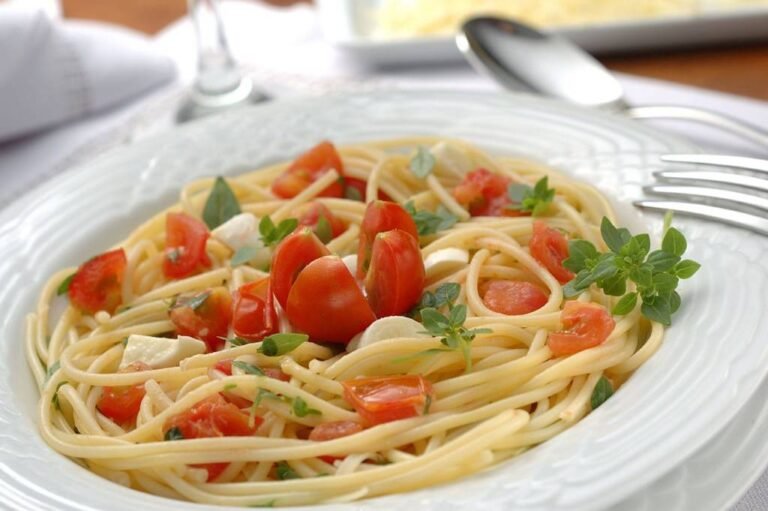 prato de espagueti com tomates e temperos, mesa posta desfocada ao redor com talheres, taças, travessa e guardanapo