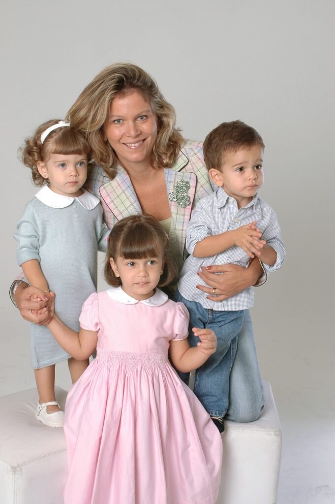 retrato familiar, mulher com 3 filhos pequenos ao seu redor, os dois menores nas suas laterais sobre puffs branco e a criança maior á sua frente, ambiente claro