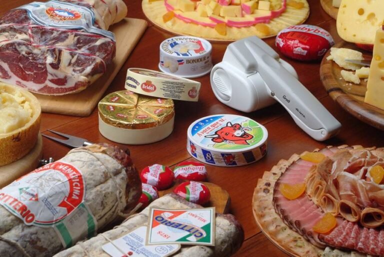 queijos inteiros e em pedaços, frios, peça de carne, salames e alguns utensílios sobre mesa de madeira
