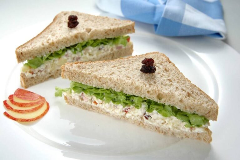 prato com sanduíche de patê com alface, com uvas passas espetadas sobre o pão de forma, guardanapo xadrez azul desfocado no fundo, fatias de maçã na lateral do prato